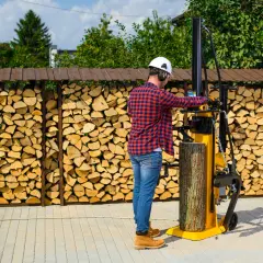Mężczyzna w kasku i słuchawkach obsługuje żółtą, poziomą łuparkę do drewna, przygotowując drewno opałowe.