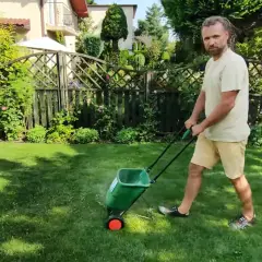 Mężczyzna używa siewnika do piaskowania trawnika, rozsypując nawóz na zielonej murawie w słoneczny dzień.