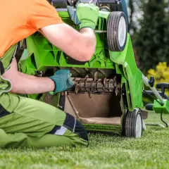 Mężczyzna w zielonym ogrodniczkach i pomarańczowej koszulce sprawdza mechanizm skaryfikatora do trawy.