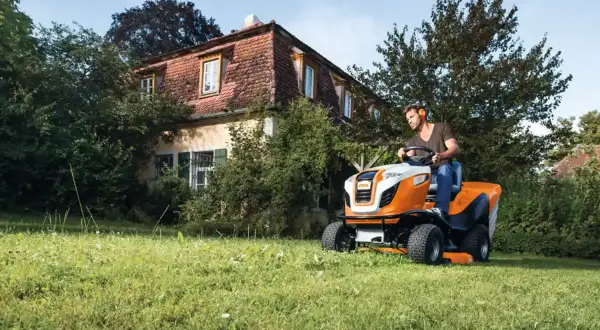 Mężczyzna na pomarańczowo-białej kosiarce samojezdnej STIHL radzi sobie z nierównym terenem. Idealny traktorek kosiarka na nierówny teren.