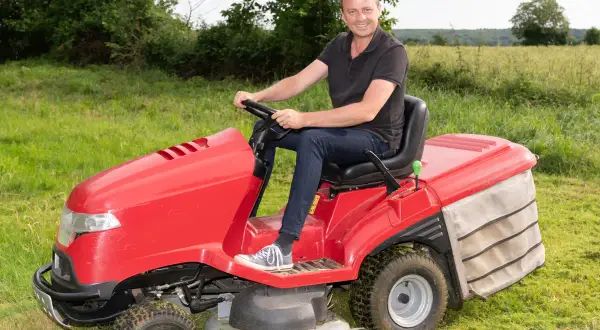Uśmiechnięty mężczyzna siedzi za kierownicą czerwonego traktorka do koszenia trawy, gotowy do pracy w ogrodzie.