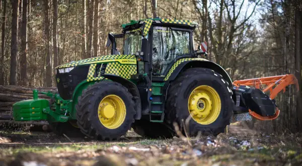 Traktor John Deere z charakterystyczną kratką w żółto-zielone wzory, wyposażony w mulczer do traktorka, pracuje w lesie.