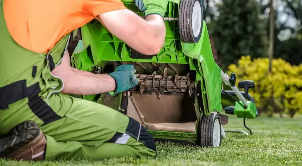 Mężczyzna w zielonym ogrodniczkach i pomarańczowej koszulce sprawdza mechanizm skaryfikatora do trawy.