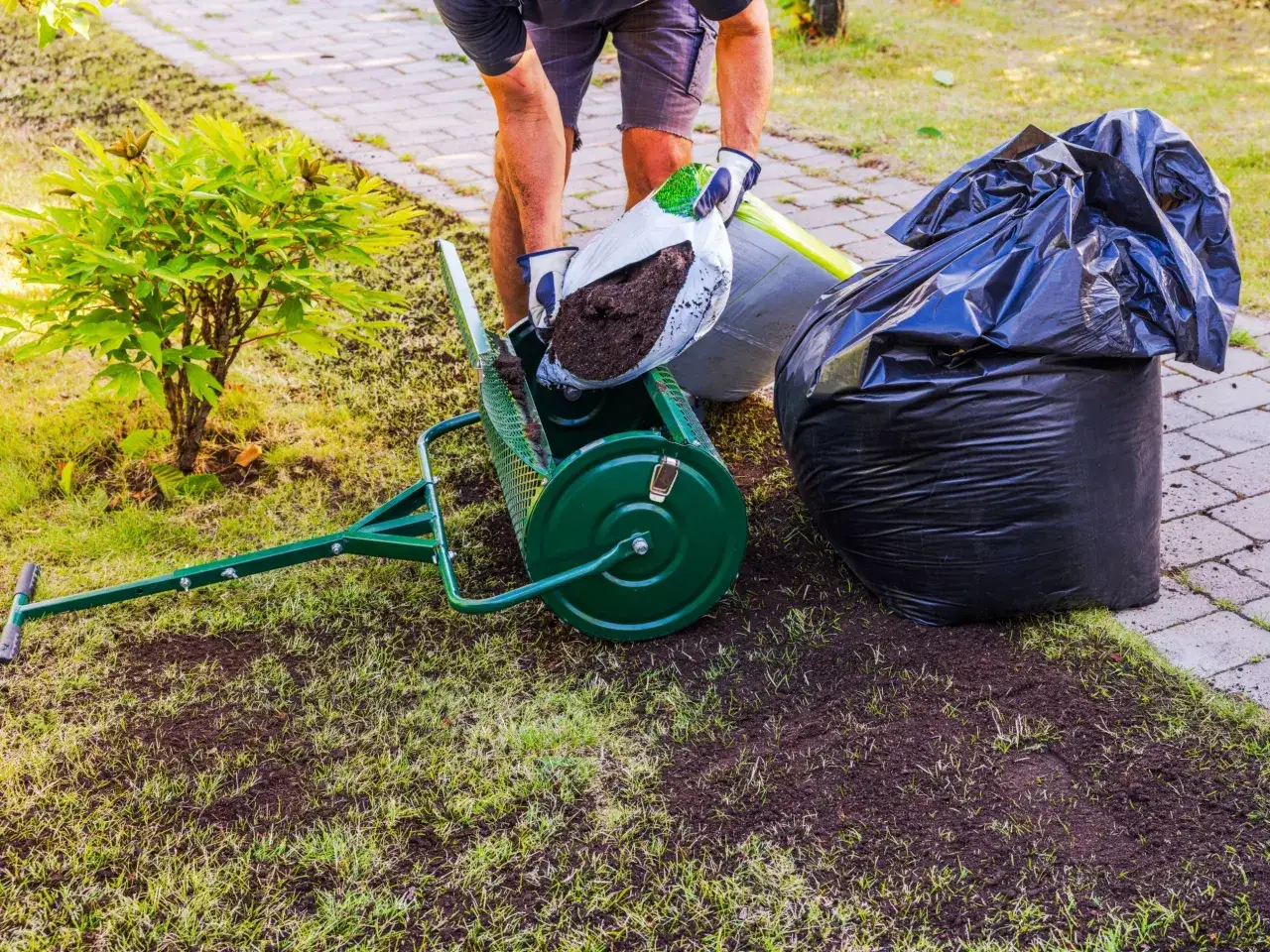 Mężczyzna wsypuje ziemię do aeratora, przygotowując się do piaskowania trawnika. Obok leżą worki z ziemią.